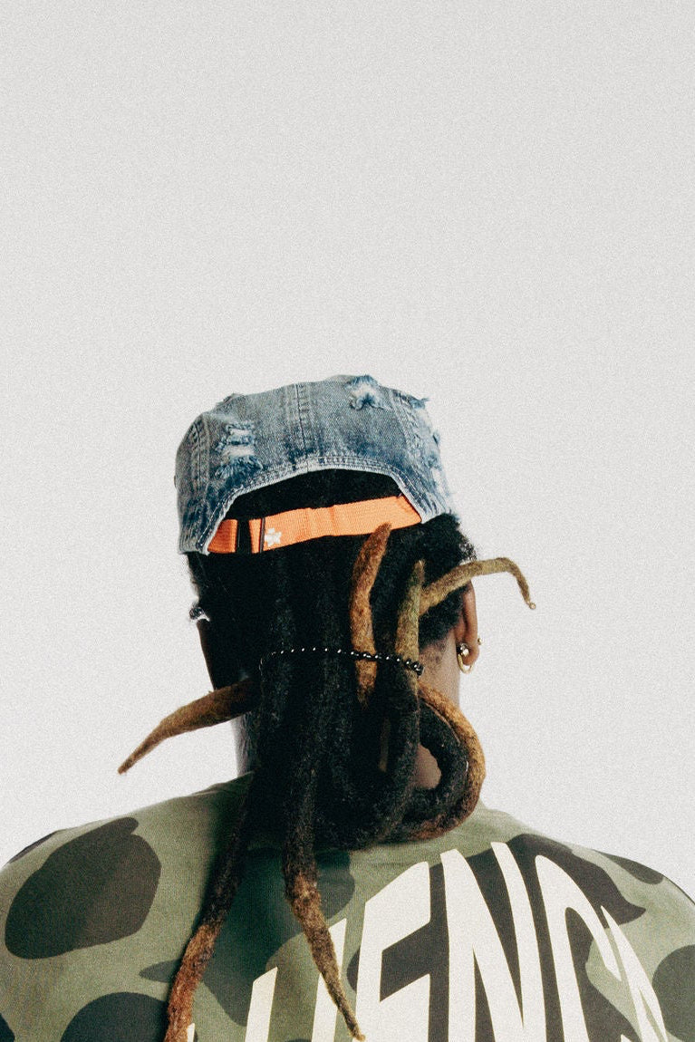 A person shown from the back wearing a green camouflage cropped jacket with large white text reading 'INFLUENCE' across the back, paired with faded dark gray jeans, a blue cap, and beige shoes, against a plain light gray studio background.