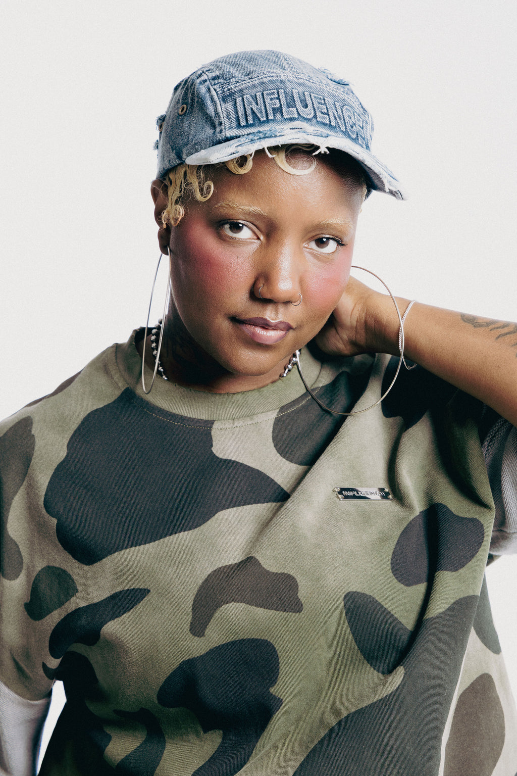 A person wearing a camouflage crewneck sweatshirt and a denim cap with 'INFLUENCE' embroidered on the front, photographed against a plain light background.