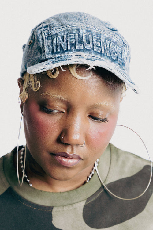 A close-up portrait of a person wearing a distressed light blue denim cap with 'INFLUENCE' embroidered on the front, paired with a green and brown camouflage crewneck sweatshirt and large hoop earrings, against a plain off-white background.