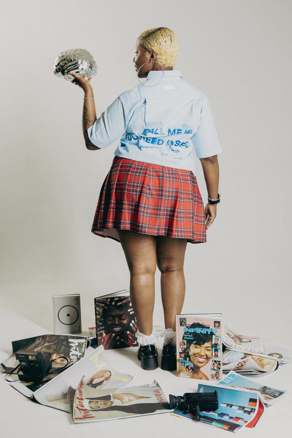 A person stands with their back to the camera wearing a light blue graphic t-shirt and a red plaid pleated skirt, holding a silver helmet. They are surrounded on the floor by scattered magazine pages and cameras against a plain off-white studio background.